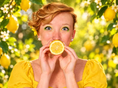 A red-haired woman in a yellow dress holds half a lemon in front of her mouth while standing among lemon branches and white blossoms.