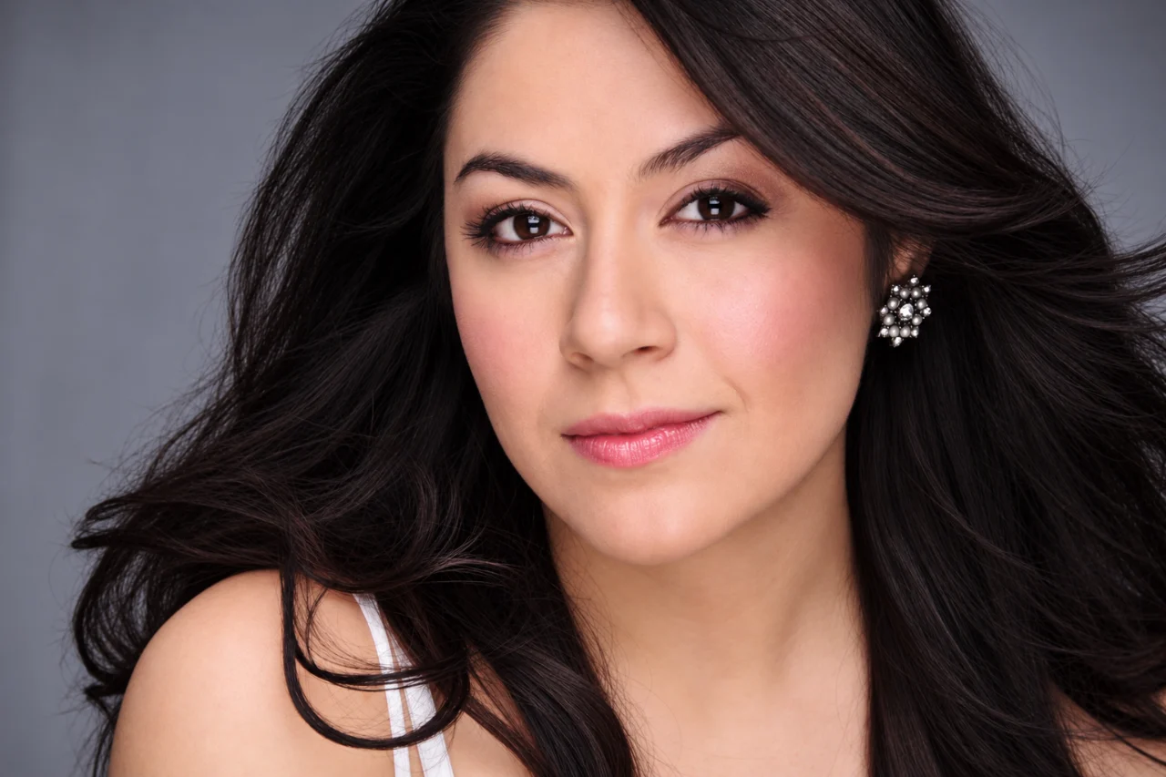 Close-up portrait of a woman with long dark hair, jeweled earrings, subtle makeup, and a soft gray background.