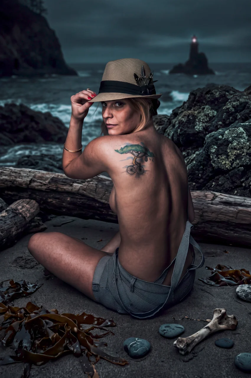 A blonde woman wearing a hat sits on a rocky beach with driftwood, crashing waves, and a distant lighthouse behind her.