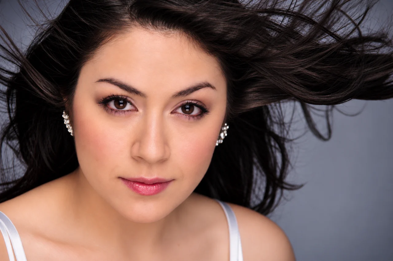 Close-up portrait of a dark-haired woman looking directly at the camera, with her hair spread around her head, jeweled earrings, soft makeup, and a pale gray background.