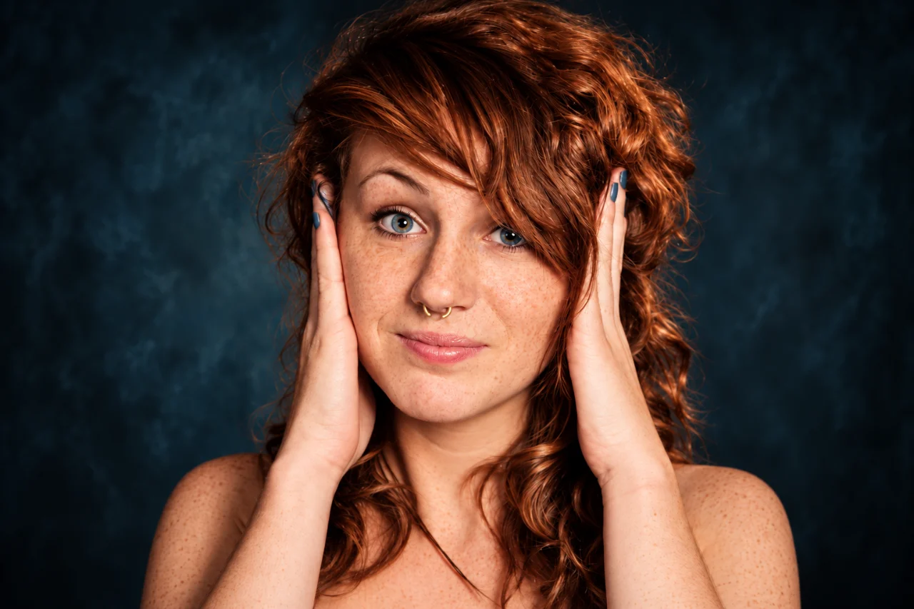 Close-up portrait of a freckled red-haired woman looking directly at the camera with both hands resting against the sides of her head. She has blue eyes, a small gold septum ring, and curly auburn hair against a dark blue background.