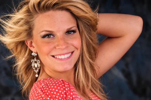 A smiling blonde woman with windswept hair wears a red polka-dot top and ornate earrings against a dark background.